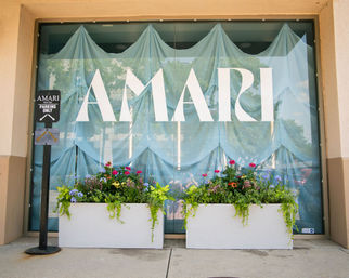 Cheerful storefront window with soft blue draped curtains, two white rectangular planters overflowing with colorful flowers and trailing greenery, and a parking sign on the sidewalk.