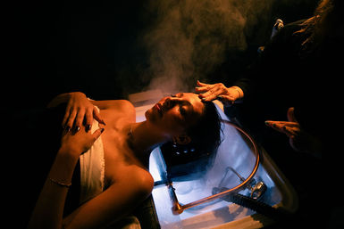 Woman reclines at a hair salon sink for a steaming spa hair treatment while a stylist massages her scalp under moody warm lighting.