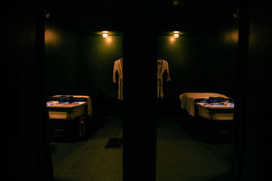 Dimly lit indoor spa treatment room with twin massage beds, illuminated trays and white robes hanging on dark walls for relaxation and massage services