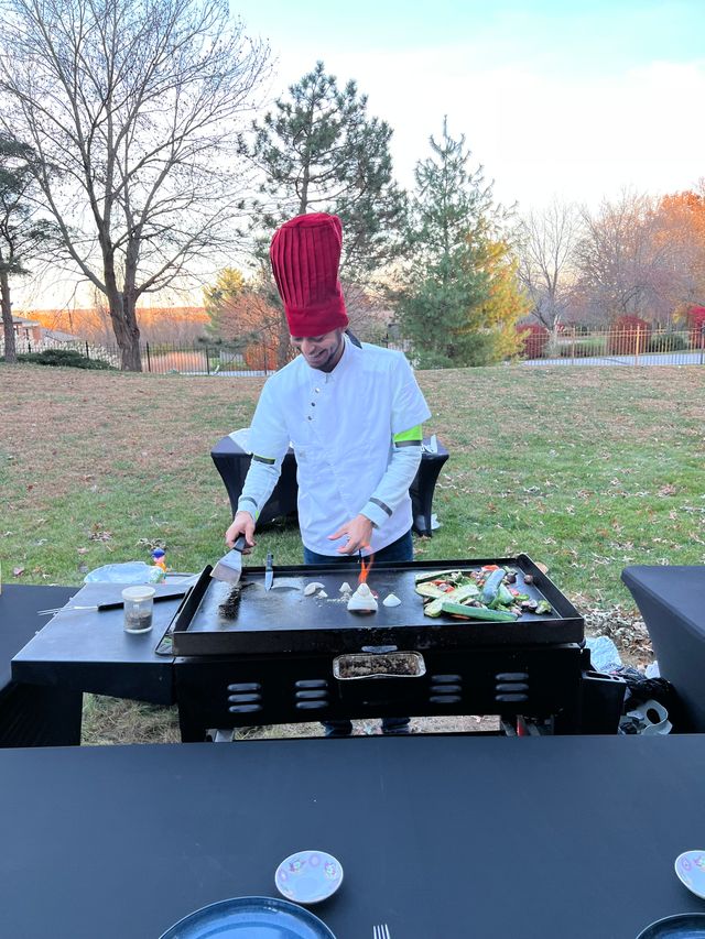 Chef in a tall red toque and white jacket cooking vegetables on a flat-top griddle outdoors in an autumn backyard, spatula in hand with a small flame on the grill and plates on a table in the foreground.