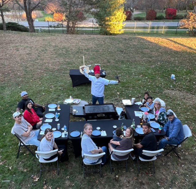 Backyard autumn family barbecue with a chef wearing a red chef hat and holding a spatula at a central griddle, surrounded by a multi-generation group seated at U-shaped black tables on the lawn with trees and a road in the background.