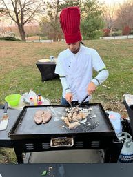 Chef wearing tall red hat and white jacket grilling steak, chicken and onions on a flat-top griddle outdoors in an autumn backyard setting