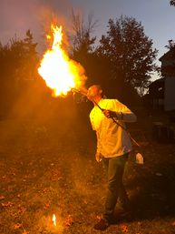Person in a white chef jacket performing fire-breathing with a long torch in a backyard at dusk, huge orange flame illuminating autumn leaves on the grass