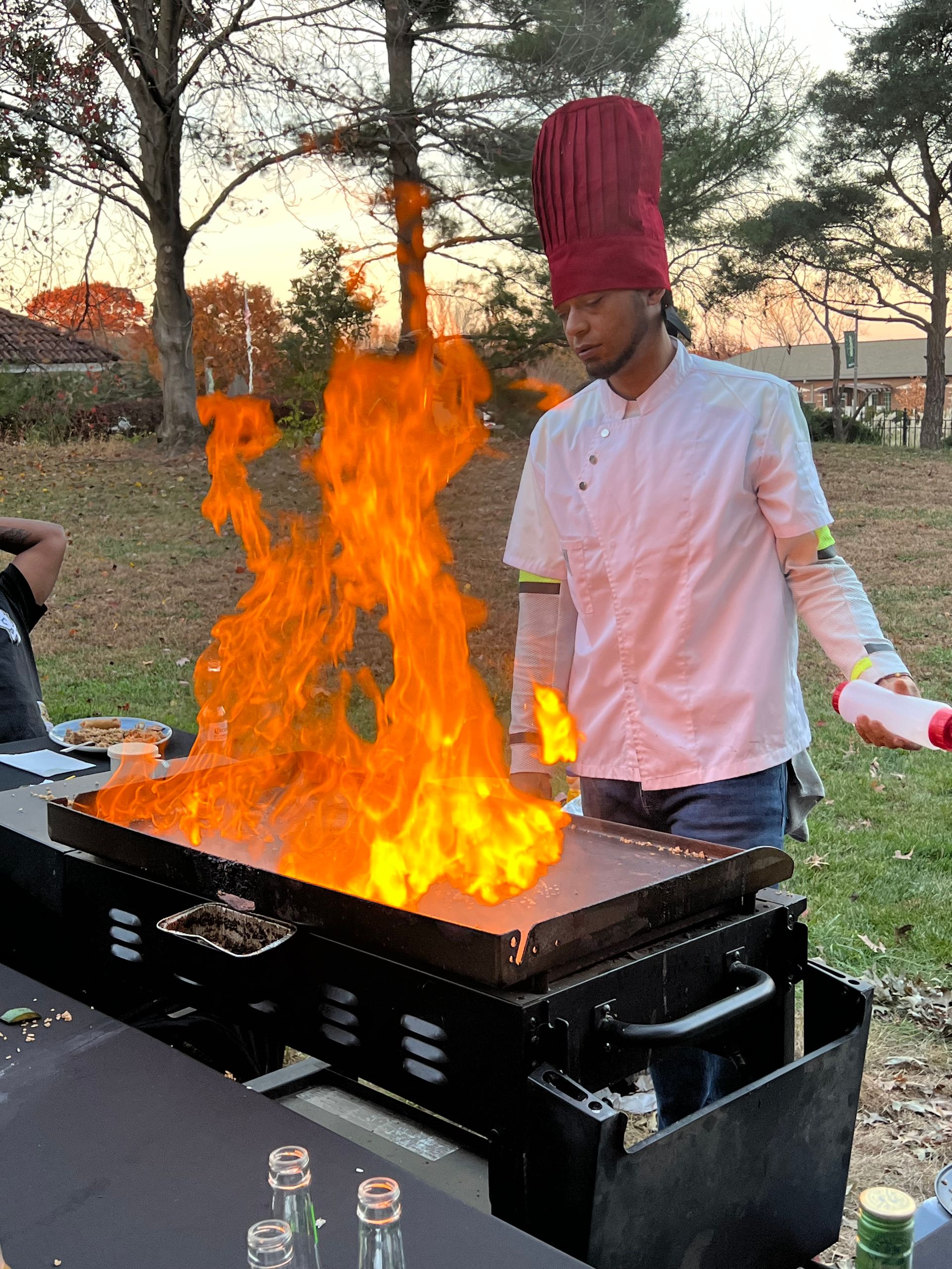 Outdoor hibachi-style chef wearing a tall red toque and white jacket cooking on a flat-top grill as towering orange flames leap up in a backyard at sunset.