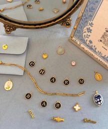 Flat lay of gold and enamel jewelry charms, a paperclip chain and initial letter discs arranged on soft blue fabric beside a vintage brass mirror and floral card.