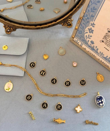 Flat lay of gold and enamel jewelry charms, a paperclip chain and initial letter discs arranged on soft blue fabric beside a vintage brass mirror and floral card.