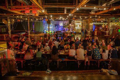 Bustling indoor warehouse-style beer hall with long communal tables, twinkling string lights, exposed wooden beams and a glowing "Rock & Roll" sign.