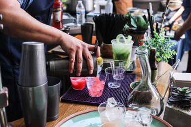 Hand pouring bright pink cocktail from metal shaker into plastic cups at a busy cocktail station with ice, shakers, herbs and a glass decanter