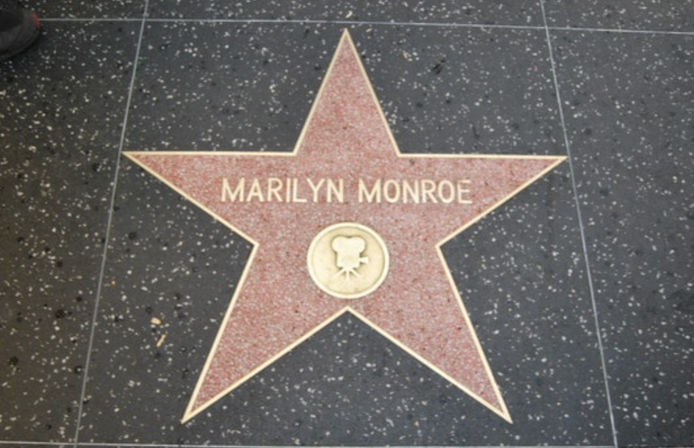Pink terrazzo Hollywood Walk of Fame star set into black speckled sidewalk, featuring a film camera emblem and an engraved celebrity name plate.