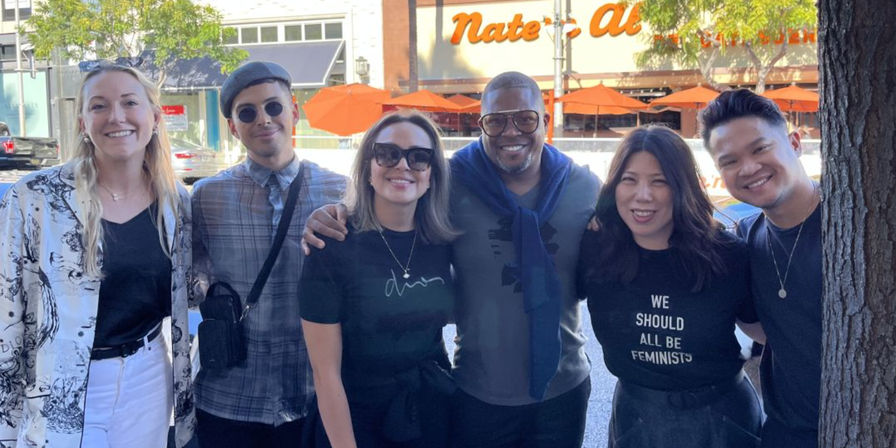 Cheerful group of six people posing arm-in-arm on a sunny downtown sidewalk outside a café with orange patio umbrellas.