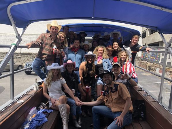 Daytime group of friends on a covered party trolley in a downtown parking lot, wearing cowboy hats, denim and boots, holding red cups and beers.