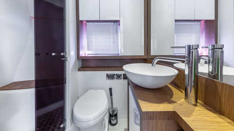 Sleek compact yacht bathroom with white vessel sink and curved chrome faucet on a wood-veneer counter, toilet next to a glass shower door, mirrored cabinets and window blinds.