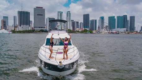 Two people in swimsuits on the bow of a white yacht cruising in front of the Miami skyline and waterfront Ferris wheel under a partly cloudy sky.