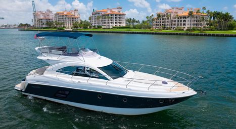 Sleek white luxury motor yacht cruising a turquoise inlet past Mediterranean-style waterfront condos and palm trees under a sunny blue sky