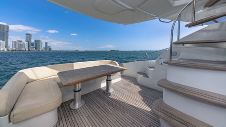 Luxury yacht aft deck with teak flooring, curved cream bench seating around a wooden table, spiral stairs, and blue bay waters with a distant city skyline under a sunny sky.