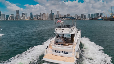 Sleek white motor yacht flying an American flag, cruising across blue-green water toward the Miami skyline under a bright, partly cloudy sky.