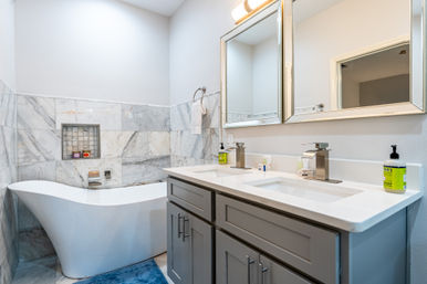 Contemporary spa-style bathroom with white freestanding tub, marble tile surround, blue bath mat, gray double-sink vanity with white quartz countertop, chrome faucets and framed mirrors.
