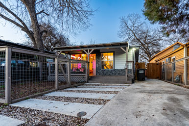 Cozy single-story bungalow with covered front porch, warm glowing interior and neon accent, fenced yard and concrete driveway framed by mature leafless trees at dusk.