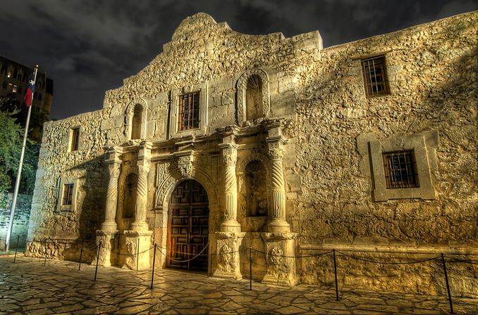 Night view of a historic limestone mission facade in San Antonio, Texas, glowing under warm lights with an ornate carved doorway and stone plaza