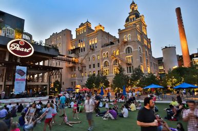 Evening scene of families and groups on a grassy urban plaza with umbrellas, in front of a warmly lit historic converted mill and tall smokestack.
