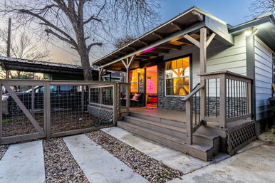 Front porch of a cozy suburban bungalow at twilight with wooden steps and railing, fenced gravel walkway, warm interior glow and a vibrant neon sign visible through the doorway.