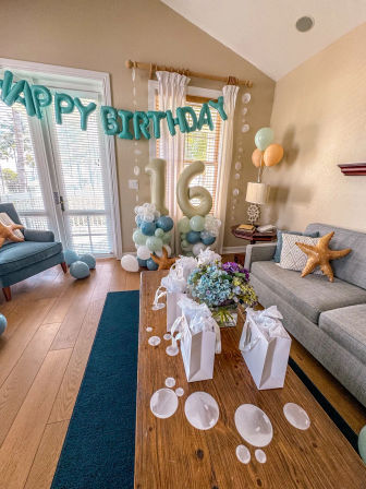 Sunlit living room decorated for a 16th birthday with teal "HAPPY BIRTHDAY" balloon banner, large gold "16" number balloons on pastel balloon columns, starfish throw pillows, white gift bags with tissue on a wooden coffee table, a hydrangea centerpiece and scattered bubble decorations.