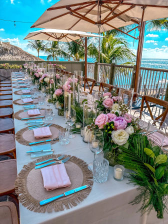 Beachfront outdoor dining on a wooden deck by the ocean, long table set for a seaside wedding with pink rose bouquets, glassware, woven chargers, pink napkins, candles, palm trees and umbrellas.
