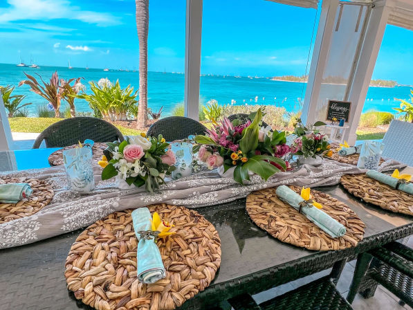 Sunny tropical ocean-view patio dining table with woven placemats, pastel napkins topped with yellow orchids and lush floral centerpieces, palm tree and sailboats on turquoise water