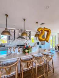 Bright modern coastal kitchen with white marble island, three brass pendant lights, four woven wooden bar stools, floral bouquet and gift bags on the counter, and large gold “27” balloons with tropical palm view through glass doors.
