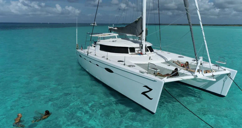 White catamaran anchored in crystal-clear turquoise tropical water, two swimmers nearby and sunbathers lounging on the bow under a partly cloudy sky.