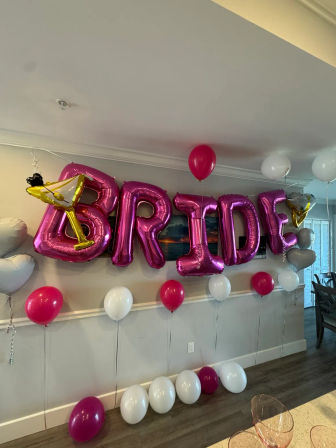 Pink metallic balloons spelling BRIDE with a gold martini-glass balloon and pink and white helium balloons decorating a home's dining area for a bachelorette/bridal shower