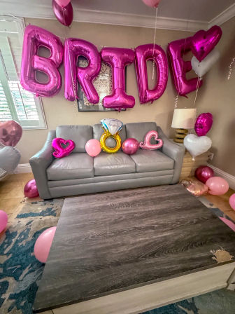 Living room decorated for a bridal shower with large metallic pink 'BRIDE' balloons over a gray sofa, gold diamond-ring and heart-shaped balloons plus scattered pink balloons on floor.