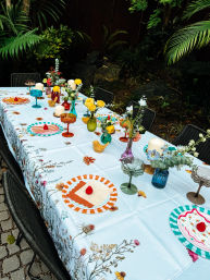Backyard garden dining table with floral tablecloth, striped plates topped with red berries, colorful glass goblets, candles and vases of yellow roses and wildflowers.