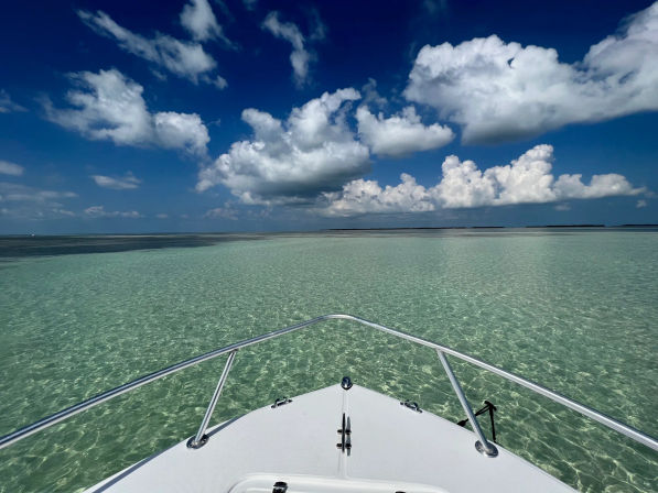 Bow of a white boat pointing into crystal-clear turquoise shallows and sandbar, with a wide horizon and a deep blue sky dotted by large fluffy white clouds