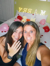 Romantic bedroom proposal: two smiling women on a bed strewn with rose petals, pink and red balloons and lit marquee letters reading 'MARRY ME', one proudly showing an engagement ring.
