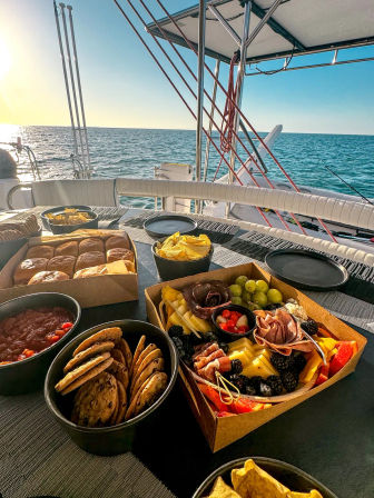 Sunlit charcuterie board with cookies and chips on a sailboat table, calm blue ocean and golden sunset on the horizon