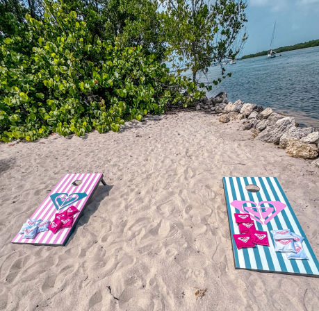 Two striped cornhole boards (pink and teal) with heart graphics and bean bags on a sandy tropical beach beside green shoreline shrubs, rocks and sailboats on calm water