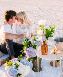 Romantic beach picnic at sunset with a couple toasting champagne on a blanket, surrounded by blue and white hydrangeas, white roses, a small table with a vase and ice bucket on sandy shore.