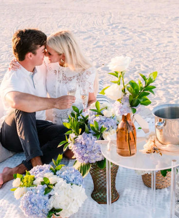 Romantic beach picnic at sunset with a couple toasting champagne on a blanket, surrounded by blue and white hydrangeas, white roses, a small table with a vase and ice bucket on sandy shore.