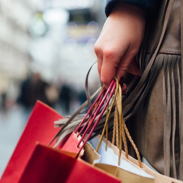 Closeup of a person's hands, holding several shopping bags, with a busy plaza in the background.