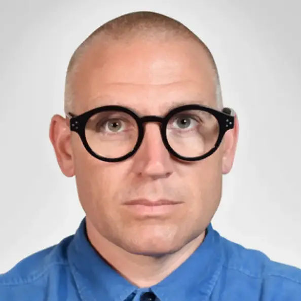 Nick Foster wearing a blue button up shirt and styish, round glasses
