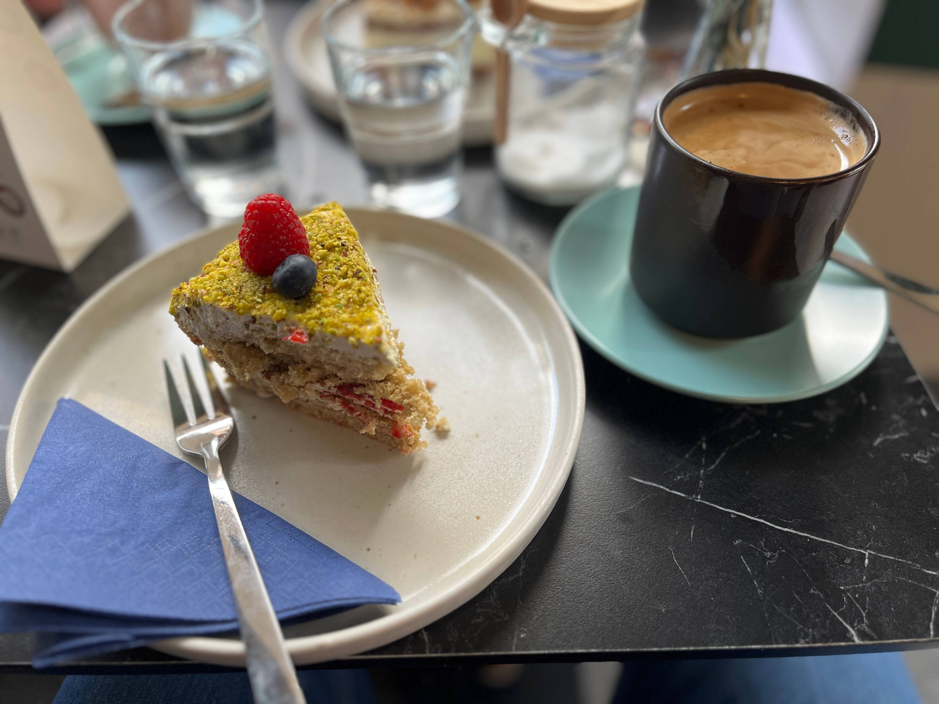 Blick auf einen Tisch, links ein weißer Telle mit einen kleinen Stückkuchen, einer blauen Servette und einer Kuchengabel. Neben dem Teller steht eine Tasse Kaffee. Im Hintergrund sieht man Wasser.