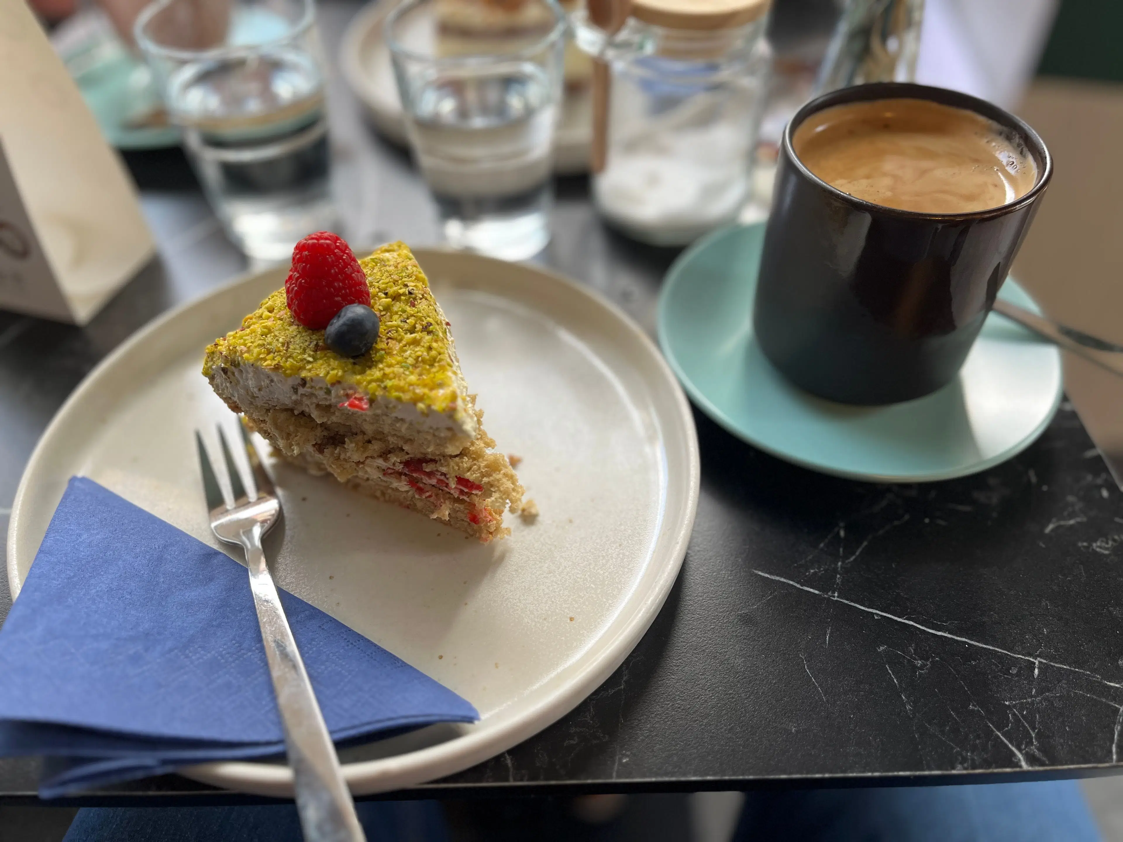 Blick auf einen Tisch, links ein weißer Telle mit einen kleinen Stückkuchen, einer blauen Servette und einer Kuchengabel. Neben dem Teller steht eine Tasse Kaffee. Im Hintergrund sieht man Wasser.