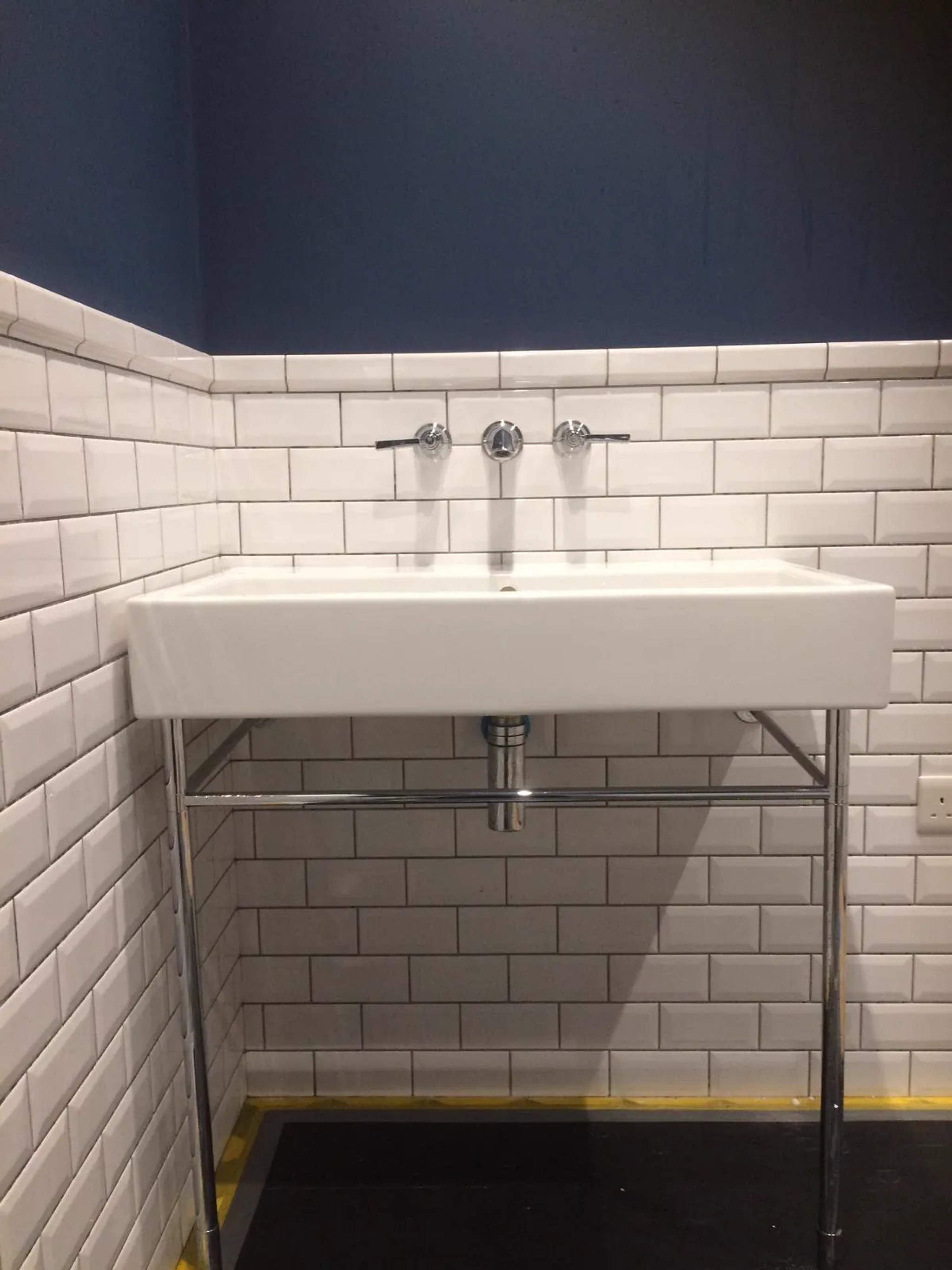 White console basin on chrome legs with wall-mounted taps and metro-tiled wall under navy paint
