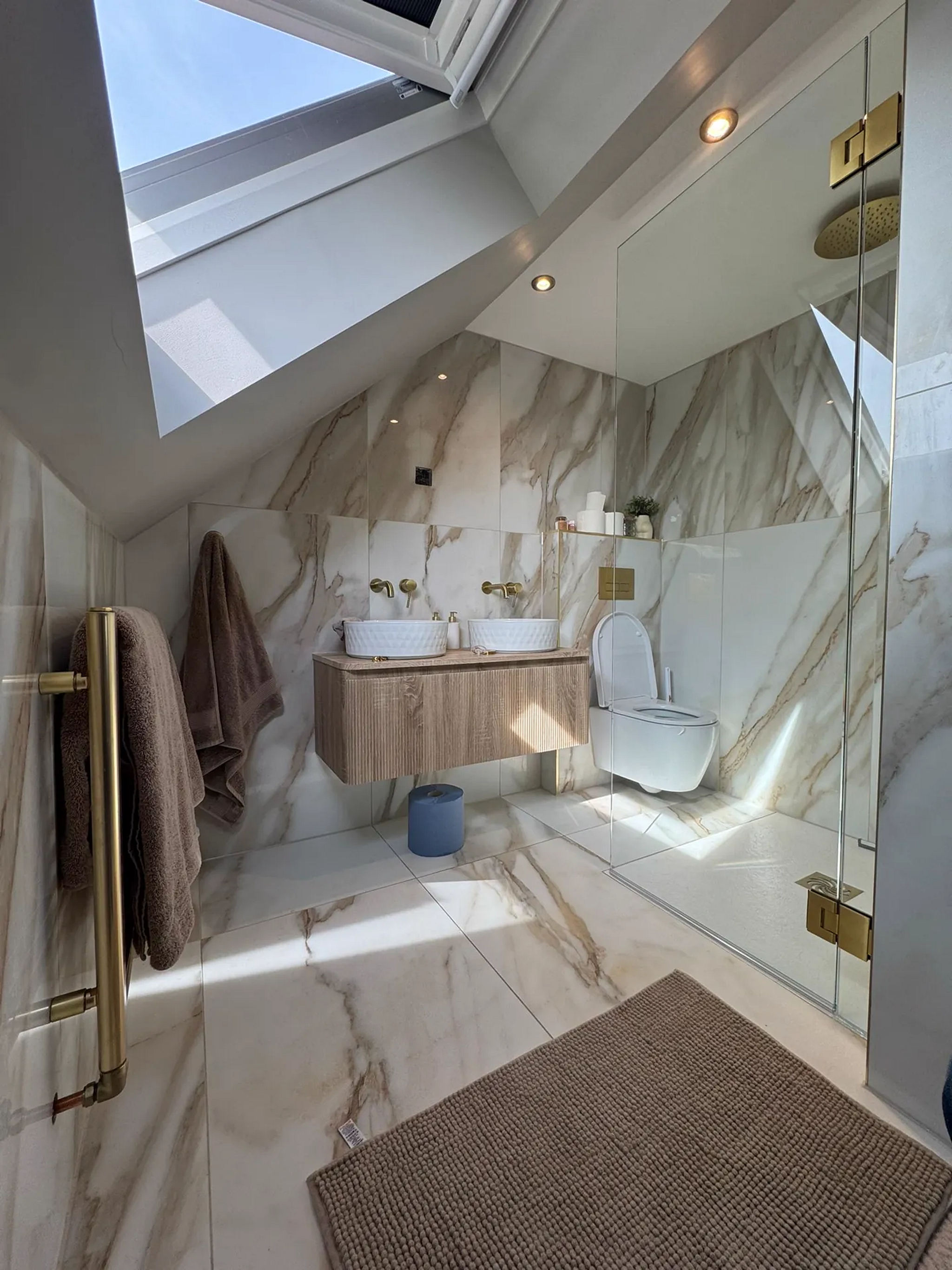 Marble loft bathroom with oak vanity, twin basins, brass towel rail and wall-hung WC under skylights
