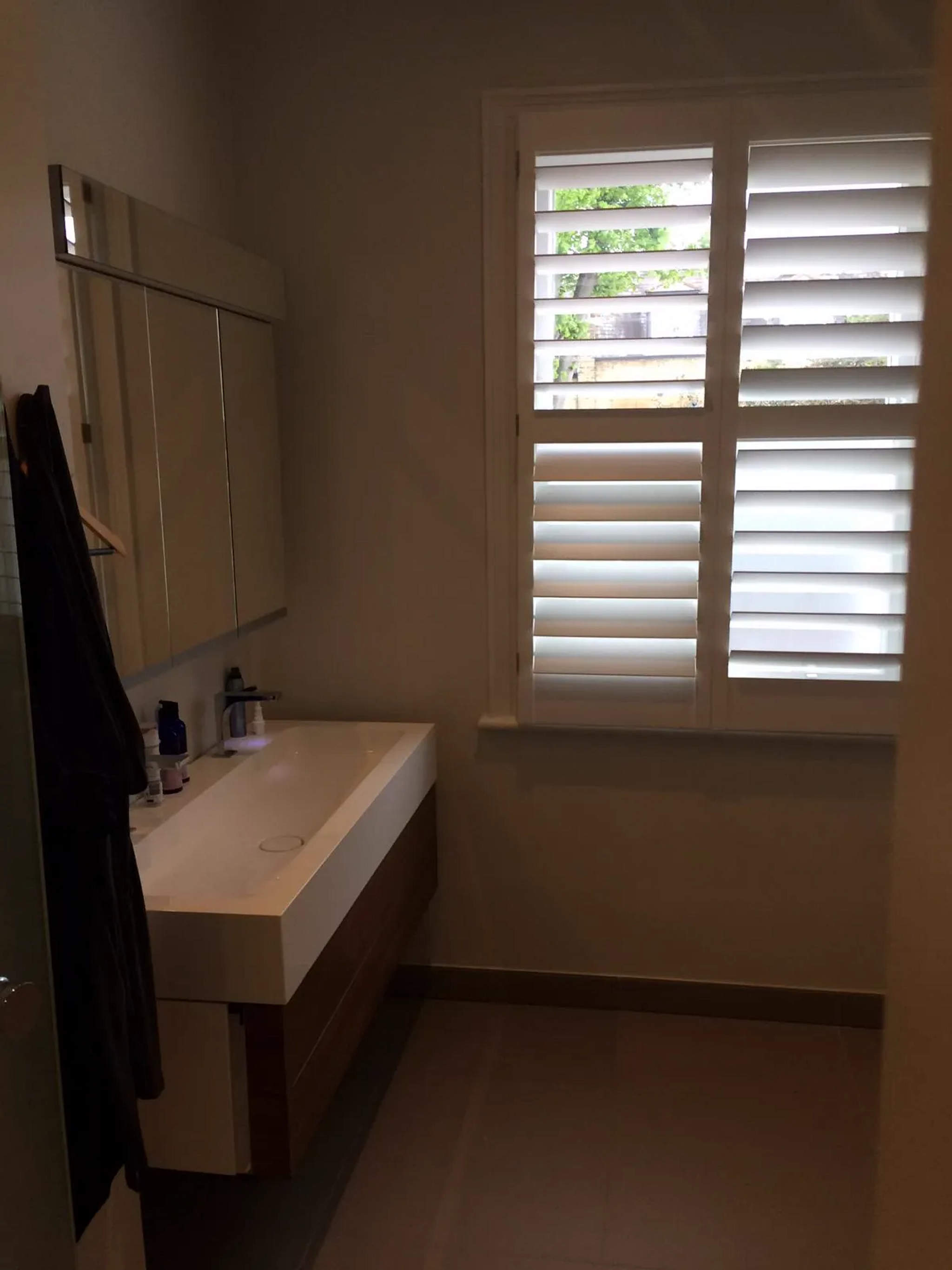 Wide white vanity basin with oak unit and mirrored cabinet beside a plantation-shutter window