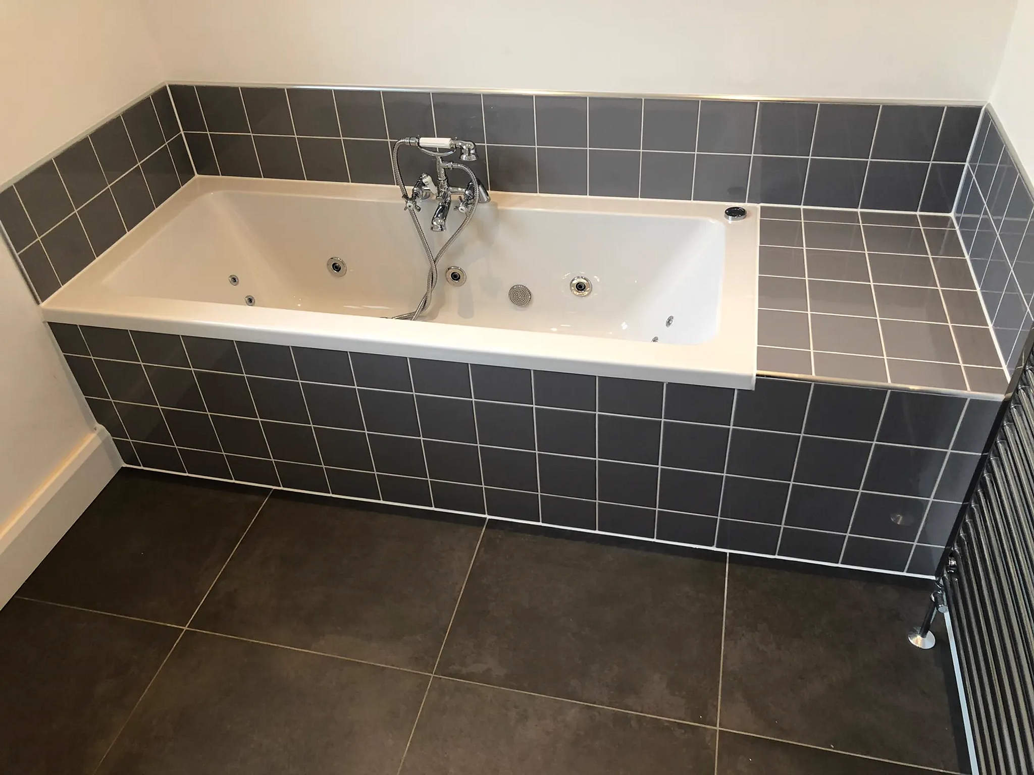 Whirlpool bath with chrome bath-shower mixer set into a grey-tiled surround and panel