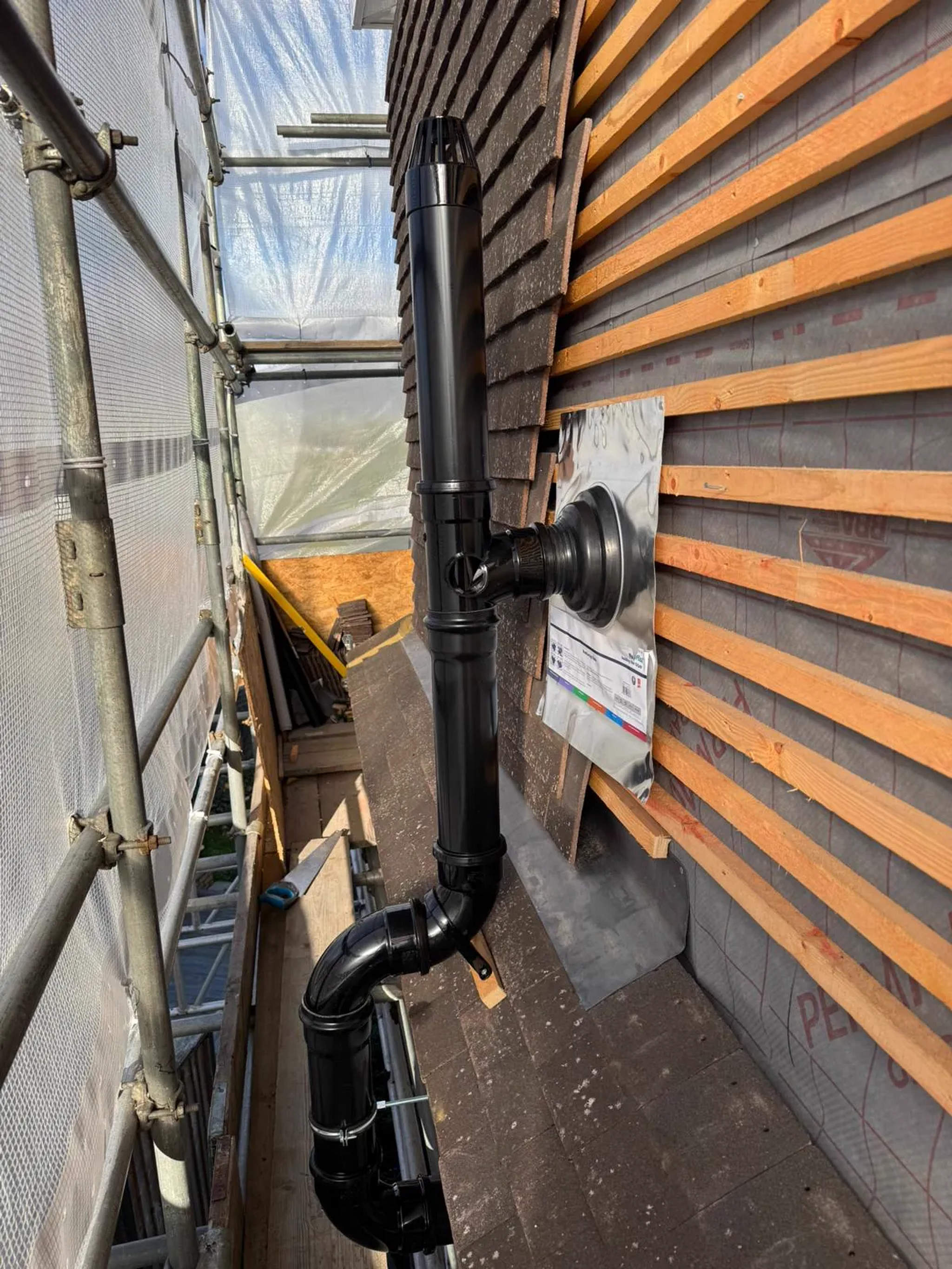 Black soil stack running up cedar-clad wall to roofline beside scaffolding on a building