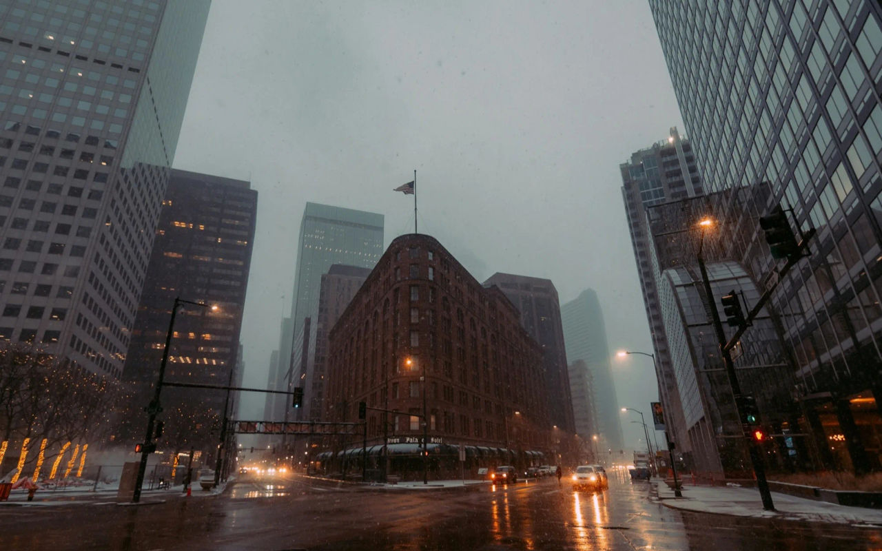 A downtown Denver intersection at dusk in the rain, a brick corner building between towers