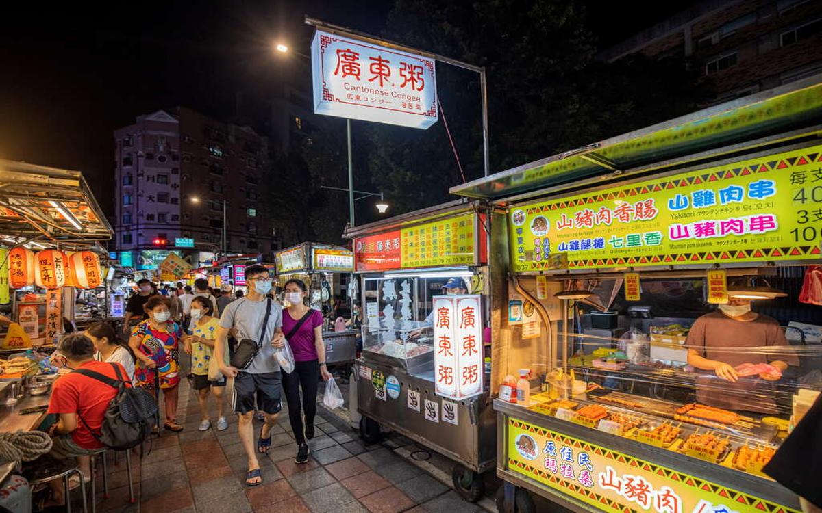 Snack stalls with colorful signs, vendors preparing their goods, and customers wearing masks moving around.